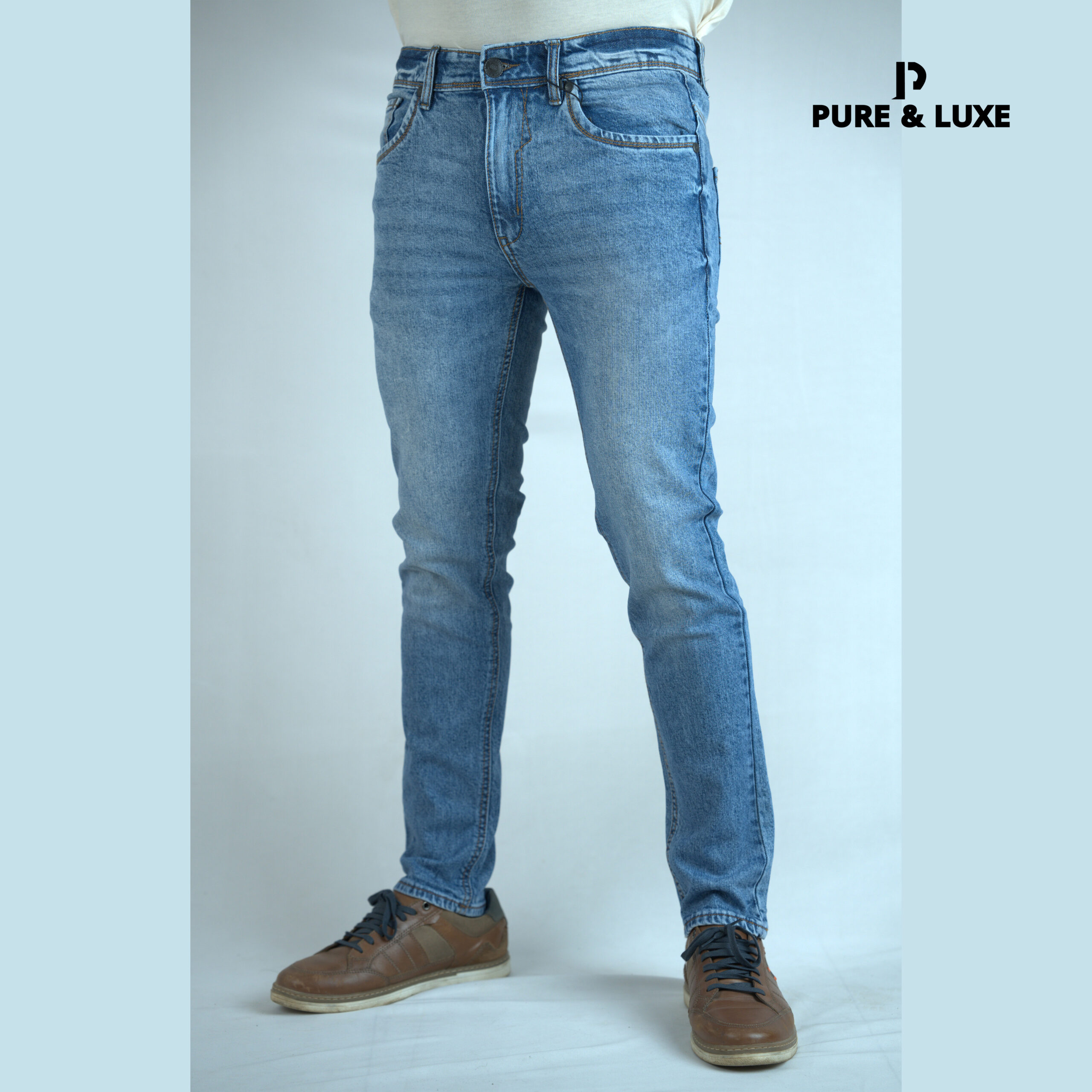 Male model wearing light blue slim fit jeans from Pure & Luxe, styled with a white shirt and brown sneakers, standing against a plain background.
