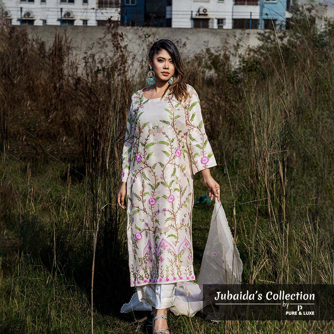 Model wearing an ivory embroidered muslin-silk 3-piece suit with pink and green floral patterns, part of Jubaida’s Collection by Pure & Luxe, standing in a natural outdoor setting.