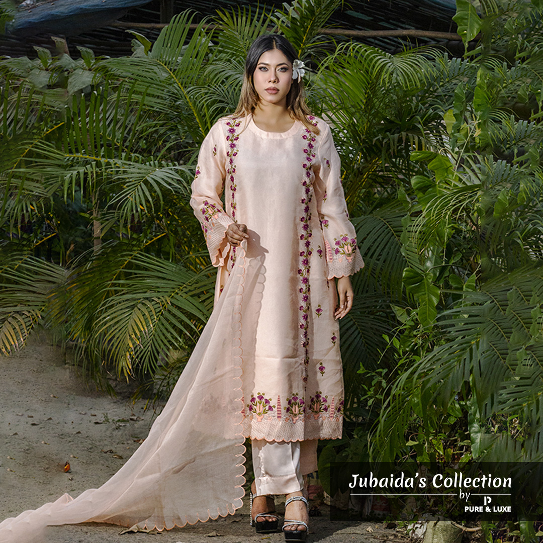 Woman wearing a blush pink embroidered muslin-silk 3-piece suit with floral detailing, holding a scalloped dupatta, standing in a garden setting.