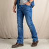 A male fashion model poses in a professional studio wearing medium-blue bootcut jeans, a heather gray crew-neck T-shirt, and an open blue denim shirt with rolled sleeves. Brown leather shoes complete the casual denim look. The main image shows a three-quarter front pose with one hand in the pocket, highlighting the jeans’ comfortable fit through the thigh and subtle flare below the knee. Three supporting panels on the right display a full front view, a full back view, and a close-up of the front pocket and waistband details. The jeans feature a medium-blue wash with natural fading, classic five-pocket construction, contrast stitching, and a bootcut silhouette designed to fit comfortably over boots. The neutral light-gray background provides a clean, modern e-commerce presentation focused on fit, fabric texture, and styling details.