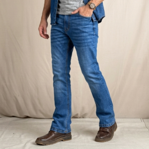 A male fashion model poses in a professional studio wearing medium-blue bootcut jeans, a heather gray crew-neck T-shirt, and an open blue denim shirt with rolled sleeves. Brown leather shoes complete the casual denim look. The main image shows a three-quarter front pose with one hand in the pocket, highlighting the jeans’ comfortable fit through the thigh and subtle flare below the knee. Three supporting panels on the right display a full front view, a full back view, and a close-up of the front pocket and waistband details. The jeans feature a medium-blue wash with natural fading, classic five-pocket construction, contrast stitching, and a bootcut silhouette designed to fit comfortably over boots. The neutral light-gray background provides a clean, modern e-commerce presentation focused on fit, fabric texture, and styling details.